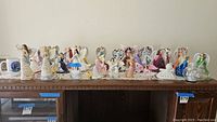 View of multiple angel figurines lined up on a wooden surface showing variety of styles, colors and sizes in group