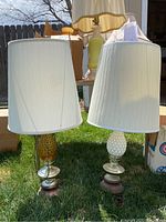 Two table lamps with pleated white fabric shades and textured glass bases in amber and white colors on metal stands, sitting outdoors on grass.