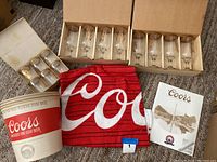 Set of boxed large stemmed Coors glasses, smaller barrel shaped glasses in box, Coors ice bucket, and red fabric Coors logo item