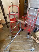Photo showing three items: red hand truck dolly with large black wheels, a red folding grocery cart wire basket, and a silver metal utility dolly with four small caster wheels on concrete floor.