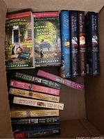 Box containing paperback mystery novels primarily by Ellery Adams with titles 'Murder in the Mystery Suite' and 'Murder in the Secret Garden' visible.