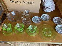 Full view of mixed glass and china items on table including green Depression glass cups and saucers, clear bowls, and decorated china plates.