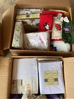 Top view of two open cardboard boxes filled with linens including new packaged sheets and assorted towels, blankets.