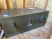 Photograph of the closed vintage olive green metal footlocker, showing riveted edges and metal clasps with some faded markings on the exterior.