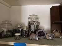 Three clear glass jars and two square glass jars containing dark tumbled stones on a shelf with agate geode halves in front