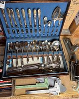 Open blue velvet-lined box containing a full set of silverware including spoons, forks, knives, and serving utensils. Visible wear and patina on utensils.