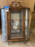 Front view of wood display cabinet with curved glass sides and decorative top design, single door with glass inset, standing on short turned legs.