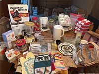 Full overview of kitchen essentials including Hamilton Beach sandwich maker and rice cooker in boxes, vintage blender, Mr. Coffee 4-cup coffee maker, kitchen textiles, baskets, plates and flatware.