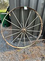 Full view of large 4 foot metal wagon wheel leaning against a metal container outdoors on gravel ground, showing weathered patina and metal spokes.