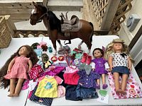 Full view of lot showing two dolls, brown plastic horse with saddle, assorted doll clothing, shoes, and small accessories laid on a white table.