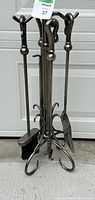 Full view of the fireplace tool set arranged on the stand showing poker, broom, shovel, and tongs in dark metal with curved handles.