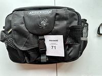 Front view of the black SpiderWire soft hip tackle box with flap and buckle closure, showing the white SpiderWire logo and spider design.