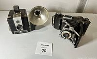 Photo showing two vintage cameras side by side, one Kodak Brownie Hawkeye Flash Model and one folding bellows camera.