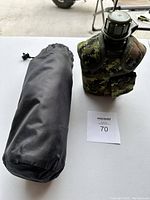 Portable inflatable pillow in black storage bag alongside canteen with camouflage case on white table