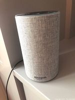 Front and side view of a single Amazon Echo 2nd Gen speaker showing light gray fabric exterior and power cord.