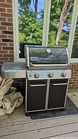 Photo showing the Weber BBQ grill closed with side shelf and cabinet doors shut. Outdoor setting with brick wall and windows in the background.