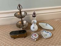 Photo showing mixed lot of porcelain and metal trinket boxes, catch-all dishes, baby powder shaker, and two-tiered metal dish stand arranged on carpet.