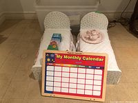 Two white wooden doll beds with quilted white and red trimmed bedding, Brain Quest card box, and magnetic monthly calendar on beds, set on beige carpet.