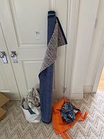 Large fabric roll standing upright against wall with smaller fabric bolts in a white bag and loose orange and blue patterned pieces on the floor.
