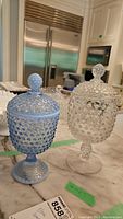 Two Fenton Glass lidded candy bowls with hobnail pattern on kitchen counter, one blue, one white opalescent.