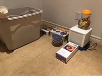 Wide shot showing plastic container, bead containers, white electronic device, and assorted boxes on carpeted floor.