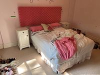 Front view of double bed with pink upholstered headboard and bedding, flanked by two white nightstands