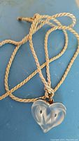 Photo of frosted glass heart-shaped Lalique pendant on beige twisted cord showing front design and metal edge trim.