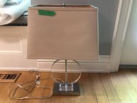 Front view of table lamp showing rectangular beige fabric shade and metallic-acrylic base with power cord.