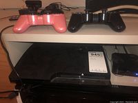 Black Sony PlayStation 3 console on shelf with two wireless controllers, one pink and one black, on top.