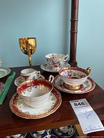 Overall view of the teacups, saucers, and gold goblet arranged on a wooden surface, showing intricate patterns and the mix of items.