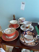 Five vintage teacups and matching saucers arranged on wood table with visible intricate patterns and gold trim.