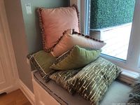 Six decorative throw cushions stacked and arranged by window, showing different styles and colors.