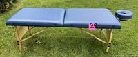 Blue portable massage table set up outdoors, showing its padded surface and wooden legs with adjustable height.