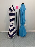Photo of two folded outdoor patio umbrellas in blue/white stripe and solid turquoise with black poles between them, set against light gray background.