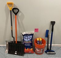 Two snow shovels, a full Blue Heat ice melt bag, a five-gallon orange bucket with partial ice melt and two cans of de-icer, and two windshield brushes with squeegee