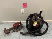 Photo showing the Shop Vac with flexible hose coiled on top and handheld vacuum cleaner with brush attachment on the left side, placed on floor against a wall.