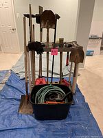 Front view of assorted yard tools including shovels, rake, pickax, hose coiled in bin, gloves, watering can, and small brooms.