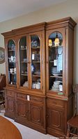 Front view of the wood buffet and hutch displaying four glass doors on the hutch and wood shelves with dishware inside. Wooden drawers and cupboards below, copper toned hardware.