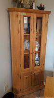 Full view of tall pine corner cabinet showing glass upper doors and wood lower doors with contents visible inside glass section.