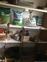 Shelf with Scotts ezseed 3 kg bag, Scotts Grass Seed Supreme 7.8 kg bag, 25 kg bag of lawn food with 16-16-16 fertilizer, and pruning shears on rack.