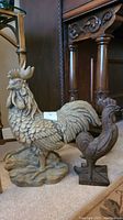 Two rooster statues side by side, larger resin with detailed feathering and smaller metal with simpler form, placed near wooden furniture.