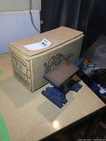 Photo of the blue 3-inch swivel base vise with small anvil on a light wood surface next to its original brown cardboard box.