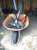 Full view of rusty metal wheelbarrow with garden tools inside including pick axe, loppers, electric weed trimmer, pruning clippers, shovel and hand hoe.