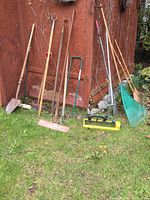 Photo of assorted yard tools leaning against a wooden shed, including shovel, edger, rakes, sprinkler, hoop hoe.