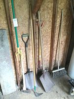 Photo showing all yard tools including shovels, sledge axe/maul, broom, fork, hoe, and garden claw leaning against a wooden wall.