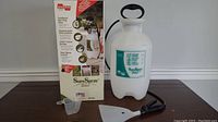 Overall product with box, sprayer tank, black hose with spray gun, and measuring cup on a flat surface