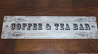 Rectangular wooden sign with 'COFFEE & TEA BAR' printed in black on a distressed whitewashed background with decorative swirls in corners.