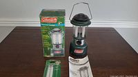 Green Coleman personal lantern next to original box, instruction manual, and extra mantles on wooden table