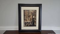 Framed picture with black frame and beige mat showing Empire State Building and street scene, viewed from a distance on a wooden surface.