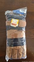 Gloves in original sealed plastic packaging, tan color with black accents and visible Carhartt logo.